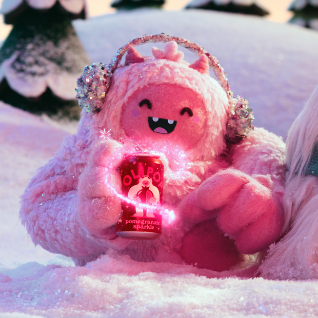 Pink plush toy with smiling face, glittery ears, holding illuminated pomegranate sparkle product in snowy setting.